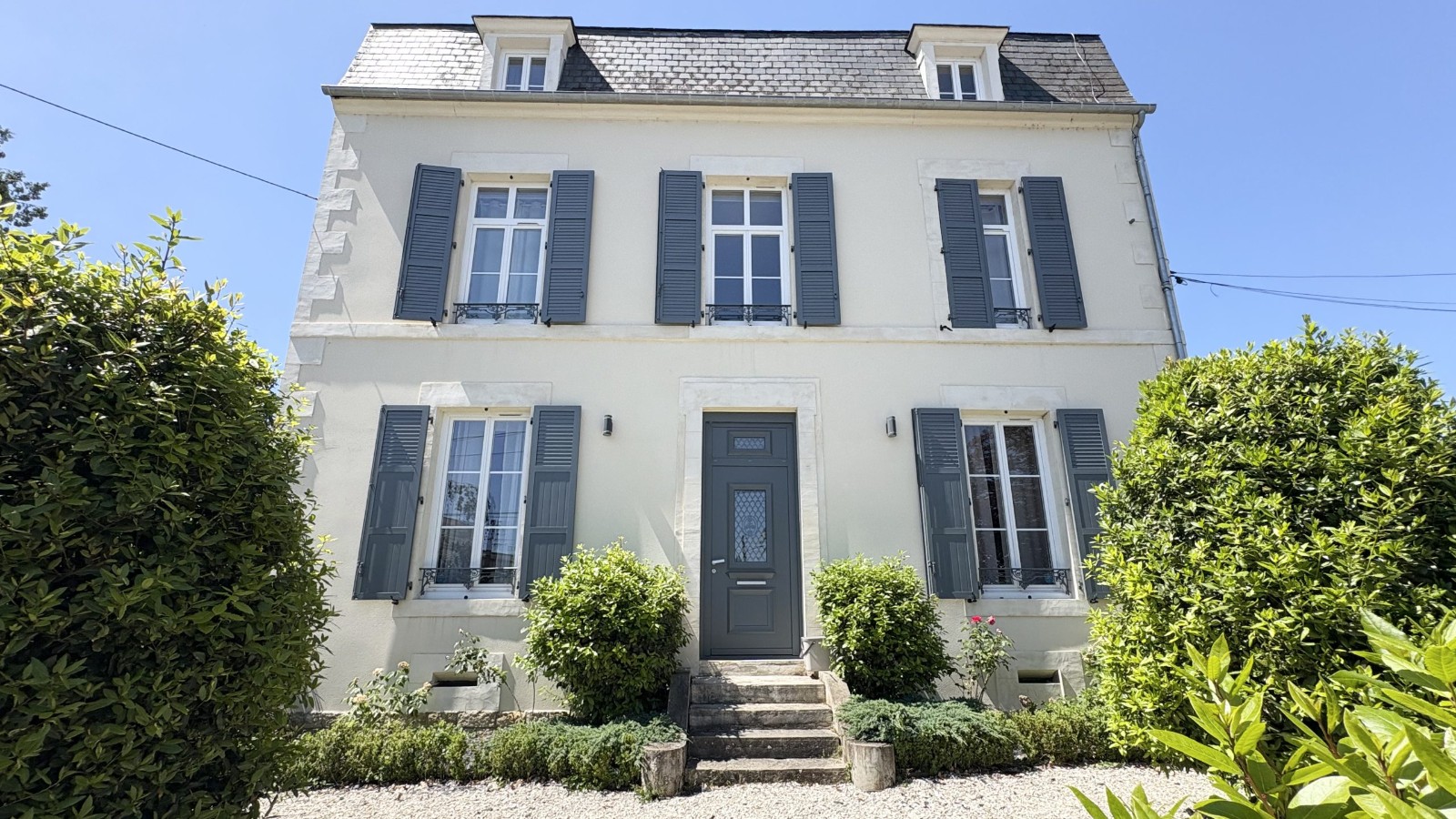 Maison de maître à vendre, façade claire aux volets bleu-gris et toit d'ardoise, avec cour en gravier et jardin arboré